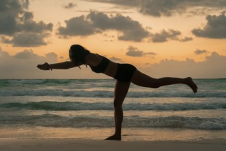 A woman balances gracefully in a yoga pose on a beach, silhouetted against a stunning sunrise. The