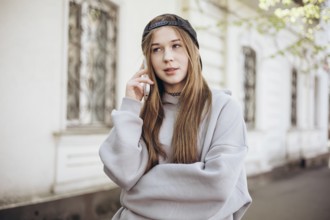 A teenage girl wearing a casual hoodie and cap is engaged in a call on her smartphone outside. She
