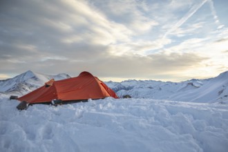 A vibrant orange tent sits atop a snow-covered peak, surrounded by majestic mountain views under a