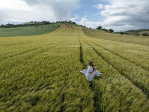 A young woman strolls through lush green fields under a wide blue sky in rural Italy, embracing the
