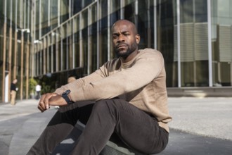 A cuban man in casual attire sits outside a modern glass building, embodying a relaxed urban vibe.