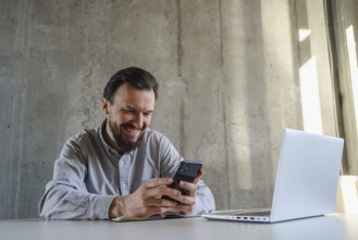 A man in a casual shirt sits at a desk, smiling while using his smartphone. Nearby, a laptop is