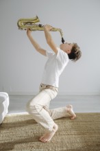 A young boy kneels inside a bright room, passionately playing a saxophone. The scene captures the
