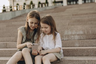 Two young girls sit together on outdoor steps in summer, engrossed in a smartphone. They share a