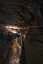 Narrow path in an illuminated cave with flowing water, gloomy adventure atmosphere, Beatus Caves,