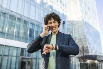 Smiling man in casual attire checks the time on his smartwatch while talking on a smartphone.