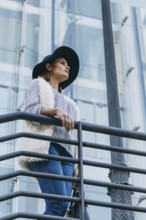 Woman in a stylish hat and fur vest stands confidently on a urban balcony. Blue jeans and a modern