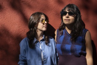 A multiethnic lesbian couple shares a moment outdoors, standing against a sunlit red wall. Both