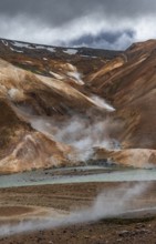 Steaming stream between colourful rhyolite mountains with snowfields, Hveradalir geothermal area,