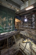 An interior construction site with exposed beams, scaffolding, and safety nets. Illuminated by