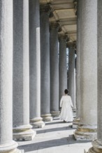 A young girl walks gracefully among towering columns, dressed for her First Communion. The sunlight