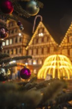 Close-up of Christmas baubles on a tree in front of a glowing structure and houses, Christmas
