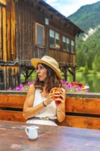 Young woman wearing a straw hat enjoying a hot drink in a mug at a wooden table with a view of lake
