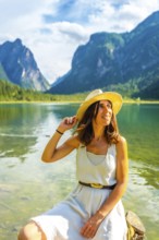 Young woman wearing a straw hat relaxing at lake dobbiaco with the dolomites in the background,