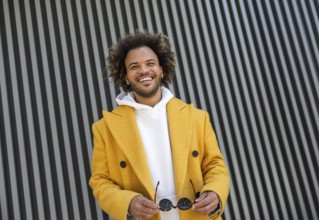 A cheerful Afro man in a yellow coat holds sunglasses, standing in front of a modern striped