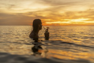 Silhouette of man relaxing in the ocean at sunset, holding a cocktail, enjoying a serene vacation