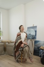 A woman sits in a bright, cozy home art studio with a colorful blanket draped over her shoulders,