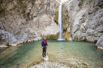 Adventurer stands on a rock in the middle of a clear stream facing a tall waterfall in Matacanes,