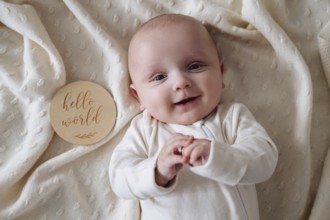A smiling baby boy lies on a soft, cream blanket. Beside him, a wooden plaque reads Hello World,