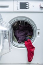 A bright red sock hangs from the door of an open front-load washing machine filled with laundry,