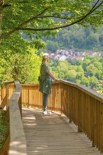 Woman on the wooden path looks at a village in the middle of a spring-like forest landscape,