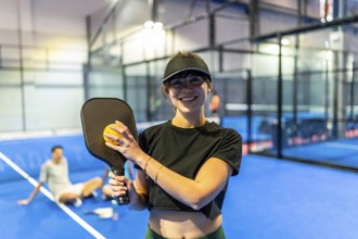 Smiling young woman holding a pickleball paddle and dimpled plastic ball, standing on a blue court