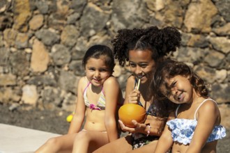 Three children in swimsuits sit happily against a stone wall. They enjoy refreshing drinks, basking