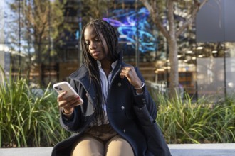 An African American businesswoman with braids intently uses a smartphone while seated outdoors on a