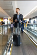 Businessman holding a smartphone and coffee while standing on a moving walkway in an airport