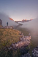 A lone hiker stands on a cliff, gazing at a stunning, mist covered valley at sunrise in the