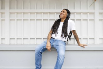 A joyful man with long dreadlocks and tattoos relaxes against a white fence, wearing headphones and