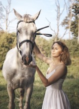 A young woman in a white dress gently touches a white horse face amidst a tranquil countryside