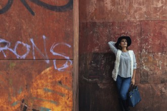 A stylish woman in casual attire with a hat leans against a graffiti-covered wall, showcasing urban