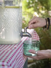 Fresh lemonade in large glass bottle with tap. Hands serving in glass. Refreshing summer drink.