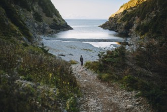 Back view of an unrecognizable man descending a rugged trail towards a serene cove in Shoe Cove