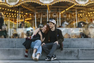 A couple sits on steps, embracing in front of a lit up carousel in Brooklyn Bridge Park. The joyful