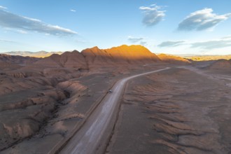 Aerial view of a vehicle in a empty desert road winding through rugged, sunlit mountains at dawn in