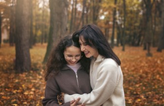 A mother and daughter share a warm embrace surrounded by autumn leaves in a forest. The cozy moment