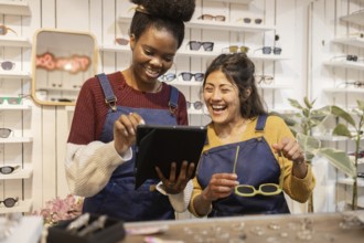Two cheerful employees in a bustling glasses shop collaborate on a digital tablet, surrounded by a