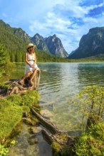 Young woman wearing a straw hat sitting on a tree trunk admiring the landscape of lake dobbiaco in