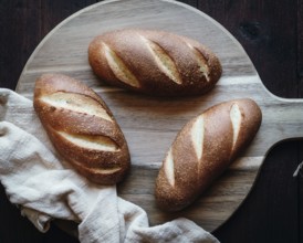 Three freshly baked loaves of artisan olive oil bread sit atop a rustic wooden board. The golden