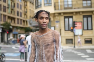 A confident man wearing a mesh top stands on a city street, embodying pride and LGBTQIA+ identity.