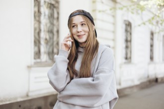 A teenage girl wearing a gray hoodie and black cap is happily speaking on her smartphone outdoors.