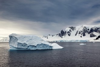 An icy mountain floats in a wide, open sea in front of a dramatic mountain landscape, icebergs in