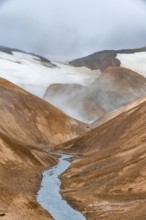 Steaming stream between colourful rhyolite mountains with snowfields, Hveradalir geothermal area,