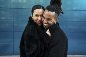 A multicultural couple stands close in a loving embrace, smiling warmly. The urban background adds