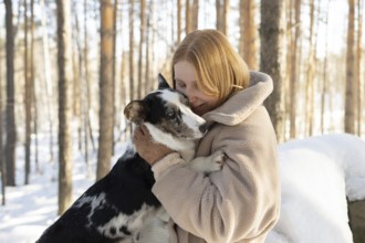 A redhead woman warmly embraces her dog, possibly a Border Collie mix, in a snowy forest setting.