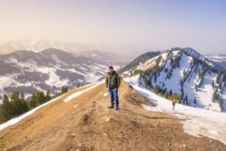 A hiker stands on a snow-covered mountain ridge with extensive mountain views under clear skies,