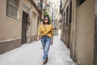 Latin woman in casual attire joyfully walks down a narrow, sunlit city street lined with old