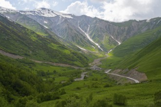 Green valley with snow-covered mountains under cloudy sky and a winding road, near Zagari Pass,